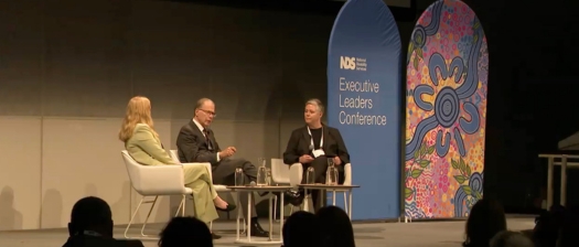 Three people sit on stage at a large conference. There is colourful event signage on stage.